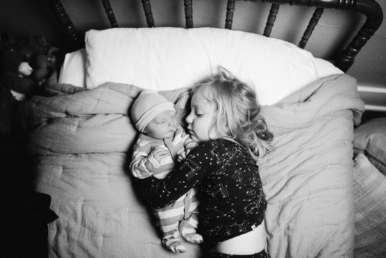 A young child lies on a bed cuddling a newborn baby who is wearing a hat and striped pajamas. Both are snuggled under a blanket, and the scene is calm and affectionate in black and white. by Justin Salem Meyer