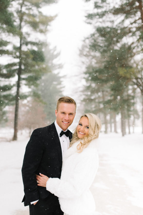 A smiling couple poses outdoors in a snowy, wooded area. The man wears a black tuxedo with a bow tie, and the woman wears a white coat. Snow falls gently around them, and pine trees are visible in the background. by Justin Salem Meyer