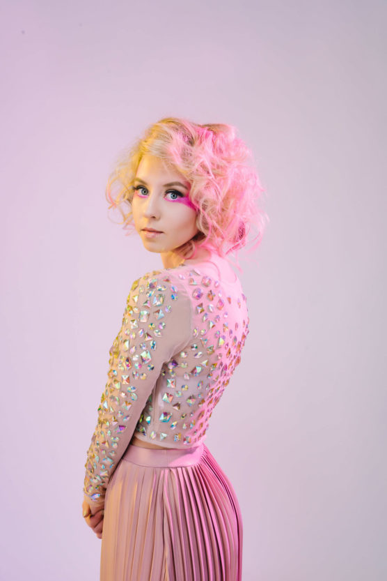 A young woman with curly blonde hair and colorful makeup gazes at the camera. She wears a light pink, long-sleeved top embellished with iridescent gems and a pleated pink skirt, standing against a soft, pastel background. by Justin Salem Meyer