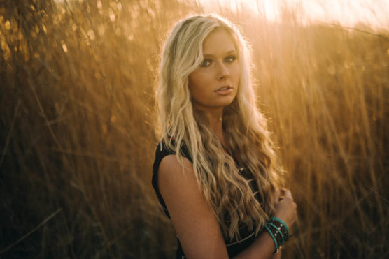 A woman with long, wavy blonde hair stands in a sunlit field of tall, golden grass, wearing a black sleeveless top and bracelets, looking slightly to the side with a calm expression. by Justin Salem Meyer