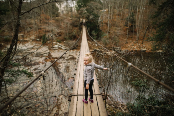 A young girl in a gray hoodie and pink shoes stands on a long wooden suspension bridge over a river, looking back toward the camera. The surrounding trees are bare, suggesting autumn or winter. by Justin Salem Meyer