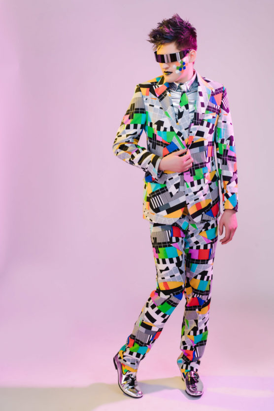 A person wearing a bold, colorful suit and matching sunglasses with a TV test pattern design stands confidently against a light pink background. They have spiked hair and metallic silver shoes. by Justin Salem Meyer
