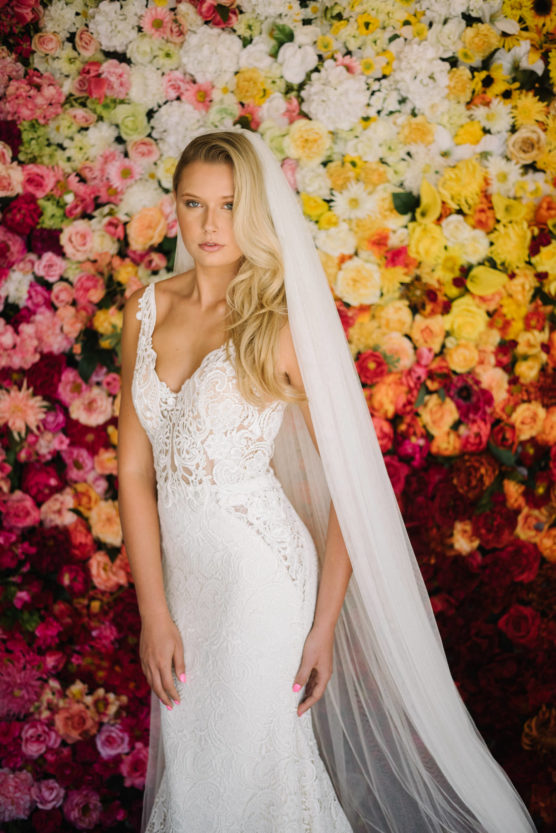 A bride in a white lace wedding dress and long veil stands in front of a vibrant, multicolored flower wall with shades of pink, yellow, red, and white blossoms. by Justin Salem Meyer