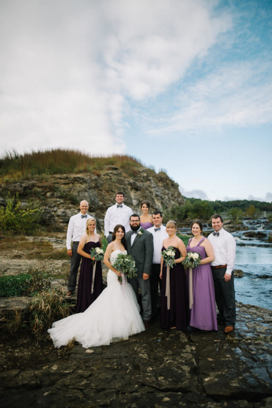 A bride and groom pose outdoors with their wedding party on a rocky riverside. Bridesmaids wear purple dresses, and groomsmen wear white shirts with dark pants. Greenery and water are in the background under a partly cloudy sky. by Justin Salem Meyer