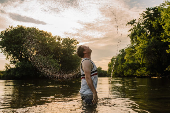 A person stands waist-deep in a river at sunset, tossing their head back and creating an arc of water droplets. Trees line the riverbank under a partly cloudy sky. by Justin Salem Meyer