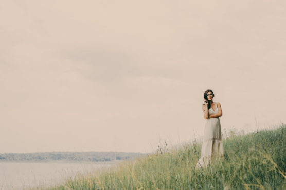 A woman in a light, sleeveless dress stands on a grassy hillside by the water, with tall grass around her and a cloudy, pastel sky above. She appears thoughtful, looking into the distance. by Justin Salem Meyer