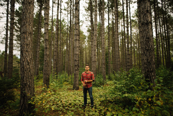 A man in a red plaid shirt and jeans stands alone among tall, straight trees in a dense forest with green undergrowth and overcast skies above. by Justin Salem Meyer