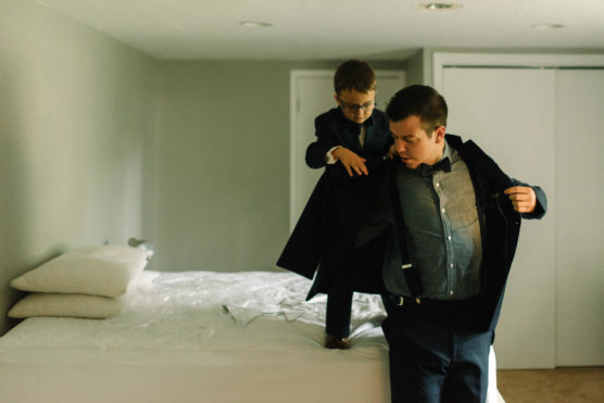 A young boy in glasses helps an older boy or young man put on a suit jacket in a softly lit bedroom with a neatly made bed and white pillows. by Justin Salem Meyer
