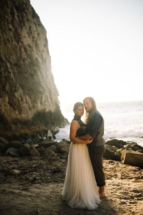 A couple stands barefoot on a sandy beach near rocky cliffs at sunset, holding each other and looking toward the camera. The woman wears a flowing white dress and the man wears rolled-up pants and a shirt. by Justin Salem Meyer
