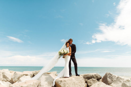 A bride and groom stand together on large rocks by the sea under a bright, partly cloudy sky. The bride wears a white dress with a long veil and holds a bouquet; the groom is dressed in a black suit. by Justin Salem Meyer