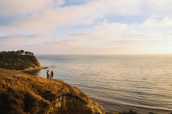 Three people stand on a cliff overlooking a calm ocean at sunset, with a rugged coastline and trees visible in the background under a partly cloudy sky. by Justin Salem Meyer