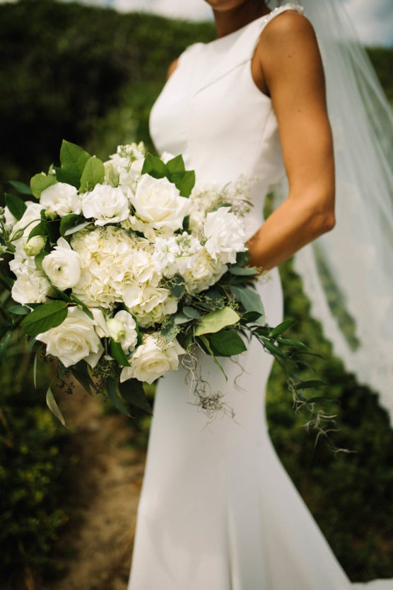 A bride in a sleeveless white gown holds a large bouquet of white roses and greenery, with a veil flowing behind her, standing outdoors. by Justin Salem Meyer