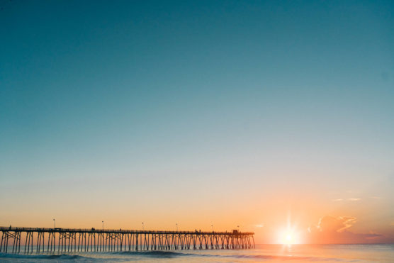A long wooden pier stretches over the ocean at sunrise or sunset, with calm waves below and a clear sky above. The sun is low on the horizon, casting a warm golden glow across the water. by Justin Salem Meyer