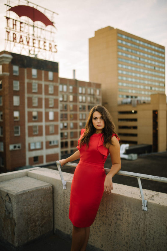 A woman in a bright red dress leans against a rooftop railing, with a cityscape of tall buildings and a Travelers sign in the background at sunset. by Justin Salem Meyer