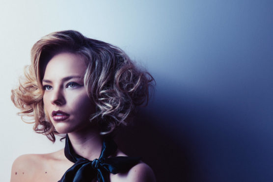 A woman with short, curly blonde hair and bold makeup looks thoughtfully to the side. She wears a dark scarf tied at her neck, and dramatic lighting casts shadows on a pale blue wall behind her. by Justin Salem Meyer