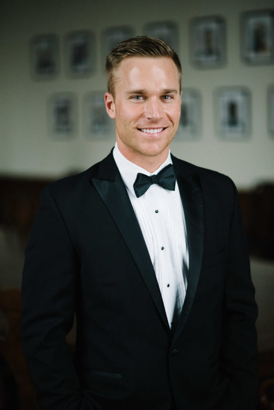 A man wearing a black tuxedo, white dress shirt, and black bow tie is smiling while standing indoors in front of a blurred background with framed pictures on the wall. by Justin Salem Meyer