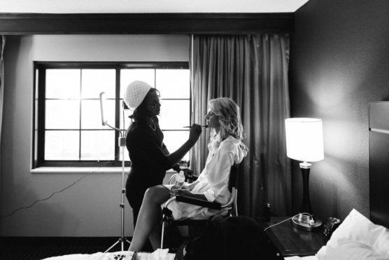 A makeup artist applies makeup to a seated woman in a well-lit room, with natural light streaming through a window and a lamp on a bedside table nearby. by Justin Salem Meyer