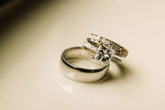 Three rings are pictured: a silver wedding band, a diamond engagement ring with a square-cut stone, and a detailed gold and silver band with small circular designs, all resting on a creamy surface. by Justin Salem Meyer