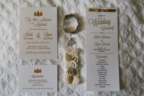 Wedding stationery set on a white quilted surface, including an invitation, information card, program with gold ribbon, and a floral wrist corsage with lace, gold leaves, and a crystal charm. Text is in gold and brown fonts. by Justin Salem Meyer