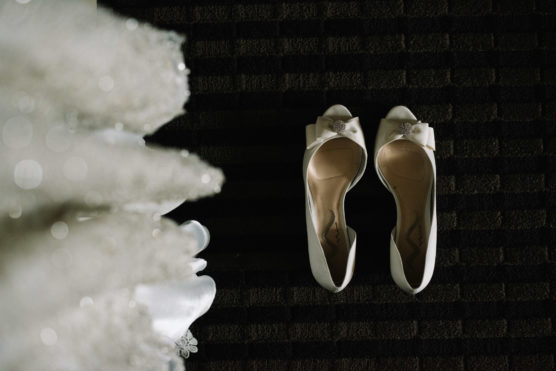 A pair of white, open-toe bridal shoes with bows sit on a dark, striped carpet next to the edge of a sparkly white wedding dress. by Justin Salem Meyer