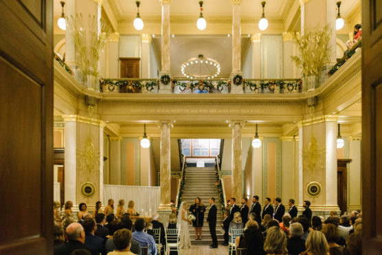 A wedding ceremony takes place in a grand, ornate hall with high ceilings, chandeliers, and a central staircase. Guests are seated while the wedding party stands at the front, facing the officiant. by Justin Salem Meyer
