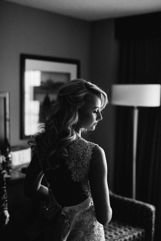 A woman in an elegant, lacy dress stands in a dimly lit room, looking to the side. Soft light falls on her face and hair, highlighting her profile. The room has a framed picture, a lamp, and a chair in the background. by Justin Salem Meyer
