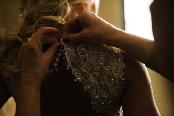 Close-up of hands fastening delicate buttons on the back of a brides ornate lace wedding dress, with softly lit blonde hair visible in the background. by Justin Salem Meyer