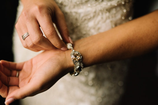 A close-up of a person’s hands fastening a sparkly bracelet on their wrist, wearing a wedding dress with lace and bead details. by Justin Salem Meyer