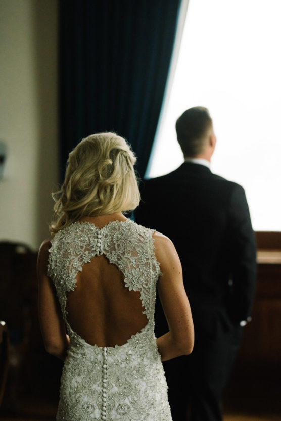 A woman in an intricate, open-back lace wedding dress stands facing a man in a dark suit, who looks out a window with blue curtains. Both are seen from behind in a softly lit room. by Justin Salem Meyer