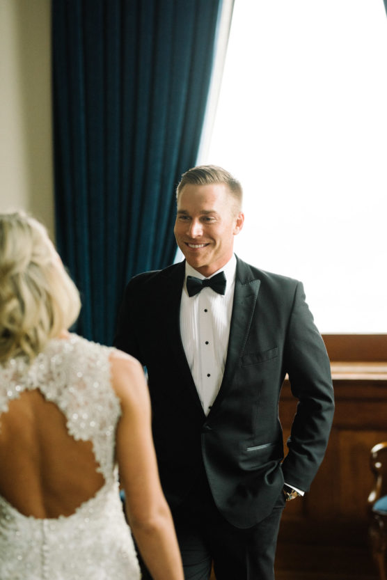 A man in a black tuxedo and bow tie smiles at a woman in a sparkly white dress with an open back, standing indoors near a large window with dark curtains. by Justin Salem Meyer