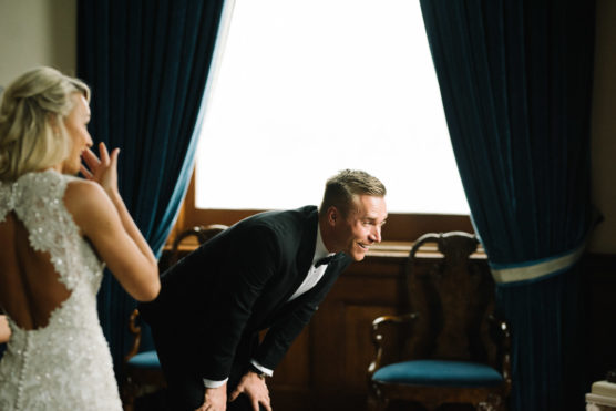 A man in a tuxedo bends forward and smiles, looking at a woman in a beaded white dress with an open back. They are indoors with dark blue curtains and antique chairs by a large window. by Justin Salem Meyer
