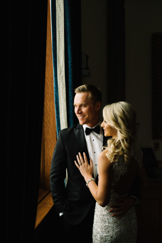 A bride and groom stand together by a window. The groom wears a black tuxedo and bow tie, smiling while looking outside. The bride, in a white beaded dress, smiles and rests her hand gently on his chest. by Justin Salem Meyer