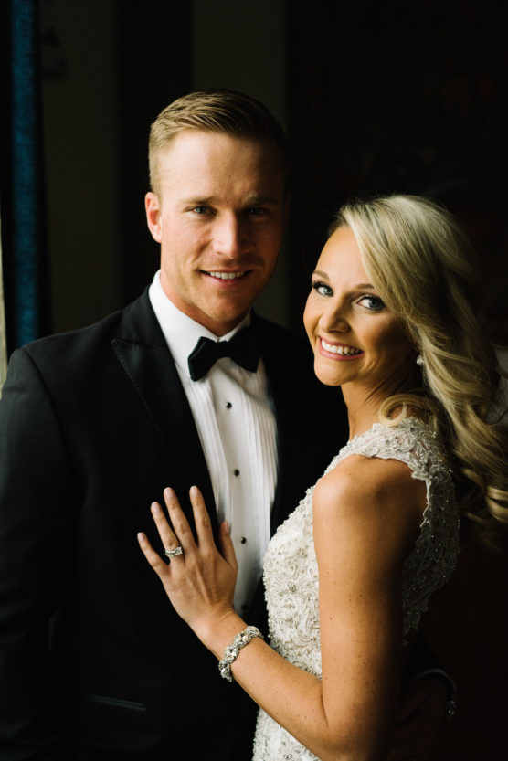 A smiling couple dressed in formal wedding attire stands close together. The man wears a black tuxedo with a bow tie, and the woman wears a detailed white gown with lace and beadwork, showing her engagement ring and bracelet. by Justin Salem Meyer
