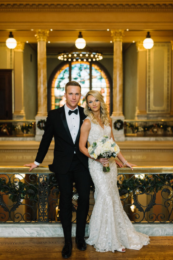 A bride in a white lace gown holding a bouquet stands beside a groom in a black tuxedo. They pose together on an ornate balcony with warm lighting and decorative marble columns in the background. by Justin Salem Meyer