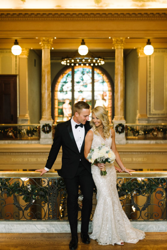 A bride in a white lace gown and groom in a black tuxedo stand close together, smiling, on an ornate balcony inside an elegant building with marble columns and a stained glass window behind them. by Justin Salem Meyer