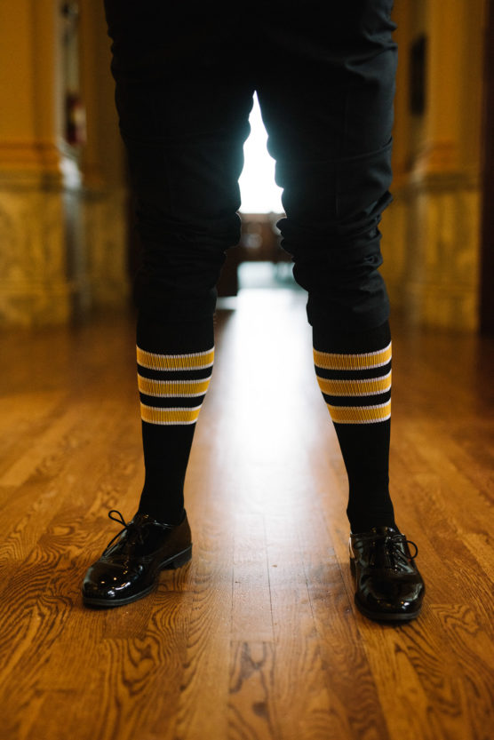 A person wearing black pants, black shiny dress shoes, and knee-high black socks with yellow and white stripes stands on a polished wooden floor in a hallway. by Justin Salem Meyer