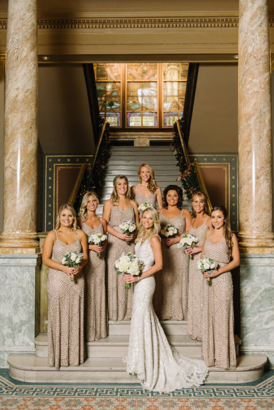 A bride in a white gown poses with seven bridesmaids in matching beige dresses, all holding white bouquets, standing on marble steps between ornate columns inside an elegant building. by Justin Salem Meyer