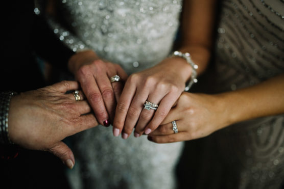 Four hands with rings and manicured nails are gently placed together, suggesting a special occasion or celebration. The background shows sequined and beaded dresses, adding a festive atmosphere. by Justin Salem Meyer