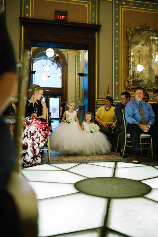 Two young flower girls in white, fluffy dresses walk down an aisle holding white bouquets, while seated wedding guests watch. Elegant decor and a large mirror are visible in the background. by Justin Salem Meyer