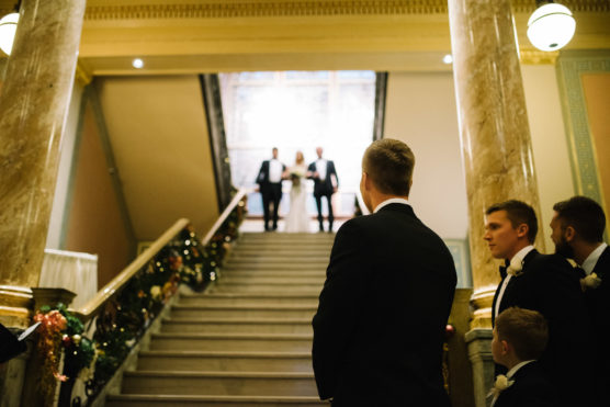 A group of men in formal suits stand at the bottom of a grand staircase, watching a bride and another person descend together at the top, in an elegant, decorated indoor setting. by Justin Salem Meyer