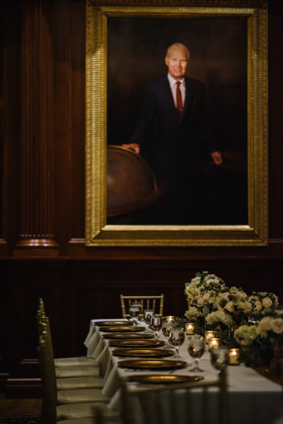 A formal dining table set with plates, glasses, and floral arrangements is positioned below a large framed portrait of a man in a suit in an elegant wood-paneled room. by Justin Salem Meyer