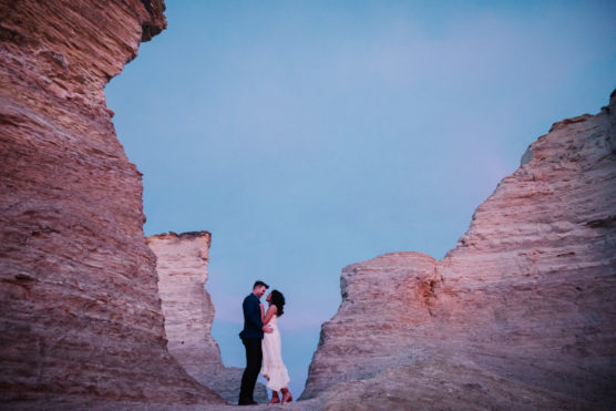 A couple stands close together in an embrace between tall, rocky cliffs at sunset, framed by the warm earth tones of the stone and a soft blue and pink sky above. by Justin Salem Meyer