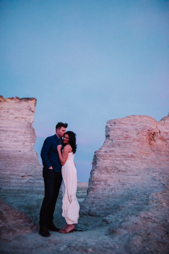 A couple stands closely together, smiling and embracing between two large rock formations at sunset, with the sky tinted pink and blue. The woman wears a white dress; the man wears dark pants and a blue shirt. by Justin Salem Meyer