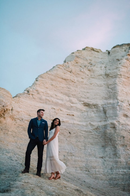 A man in a suit and a woman in a white dress stand holding hands on a rocky slope with large, pale cliffs behind them, under a pastel blue sky at sunset. by Justin Salem Meyer
