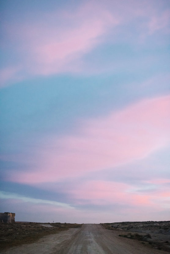 A dirt road stretches into the distance under a vast sky, filled with soft pink and blue clouds during sunset or sunrise. The landscape is flat and mostly empty. by Justin Salem Meyer