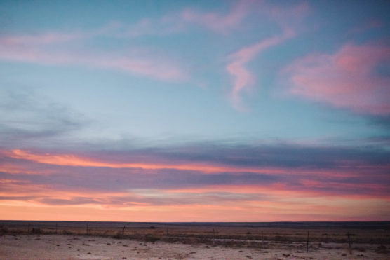 A vast, flat desert landscape under a dramatic sky at sunset, with layers of pink, purple, and blue clouds. The horizon is low, and there is sparse vegetation and a wire fence in the foreground. by Justin Salem Meyer