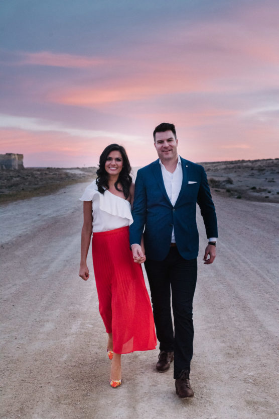 A smiling couple walks hand-in-hand down a dirt road at sunset. The woman wears a white top and long red skirt; the man wears a blue blazer, white shirt, and dark pants. The sky is pink and purple with soft clouds. by Justin Salem Meyer