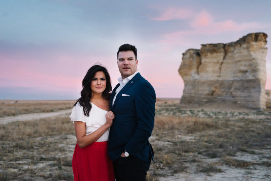 A couple stands together in a dry, open landscape at sunset. The woman wears a white top and red skirt, and the man wears a blue suit. Large rock formations and a pastel sky are visible in the background. by Justin Salem Meyer