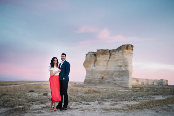 A smiling couple stands close together in a field at sunset, with large rock formations and a pink and blue sky in the background. The woman wears a red skirt and white top; the man wears a blue suit. by Justin Salem Meyer