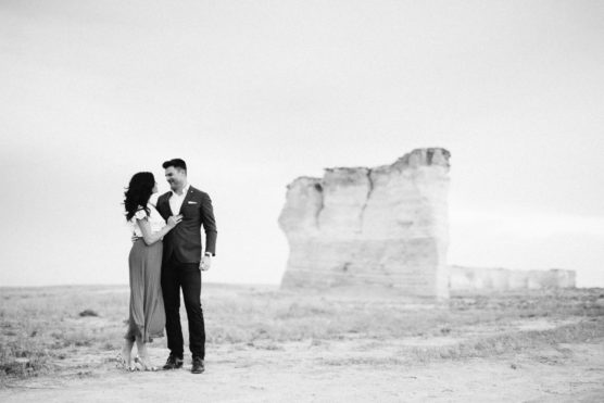 A couple stands together on a barren landscape, smiling at each other. The woman wears a dress and the man wears a suit. In the background, a large rock formation rises from the flat ground under a cloudy sky. by Justin Salem Meyer